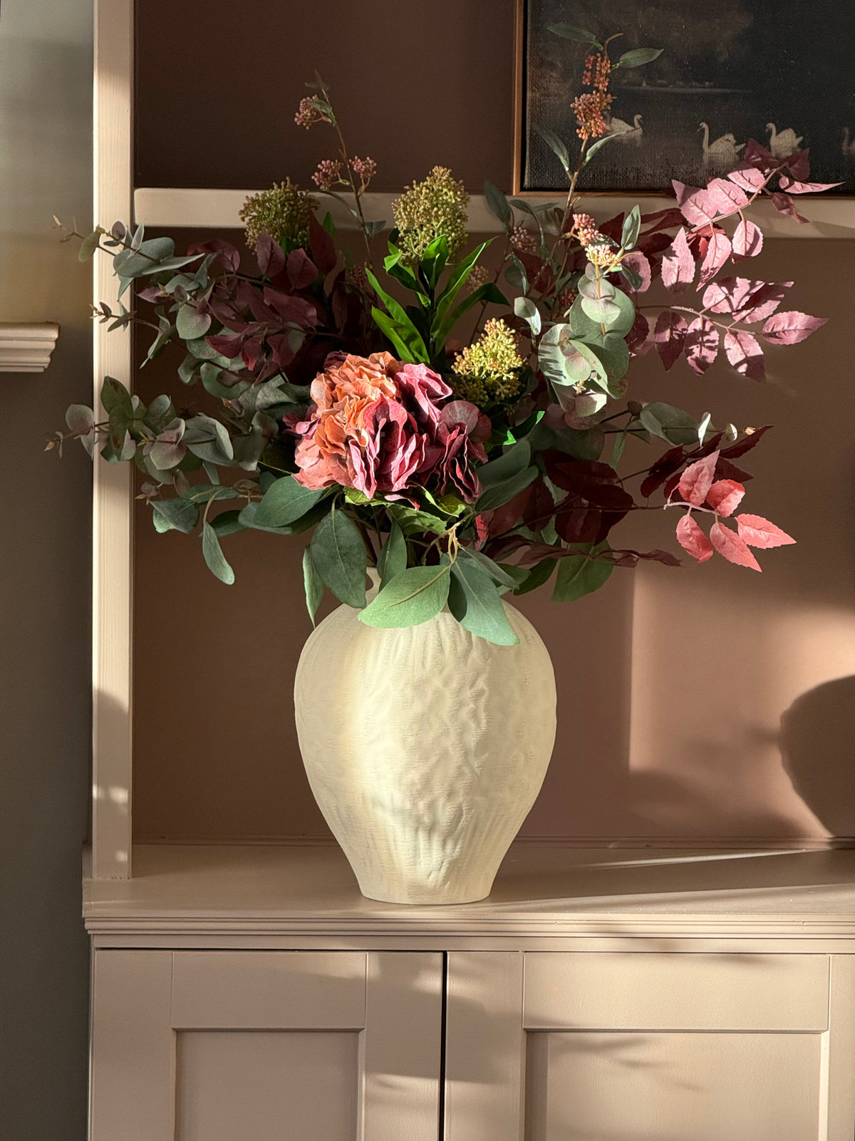 A faux floral arrangement in a textured cream ceramic vase, displayed on a light-colored wooden cabinet against a warm brown wall.