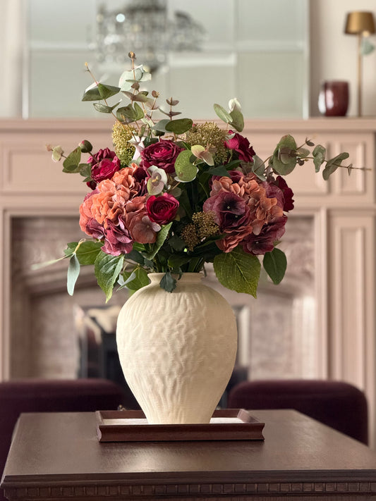 A faux floral arrangement in a textured cream ceramic vase, placed on a dark wooden table in front of a softly blurred fireplace background.