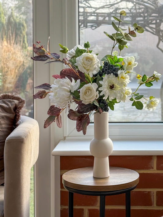 Artificial Floral arrangement in a white vase on a small round table by a window.
