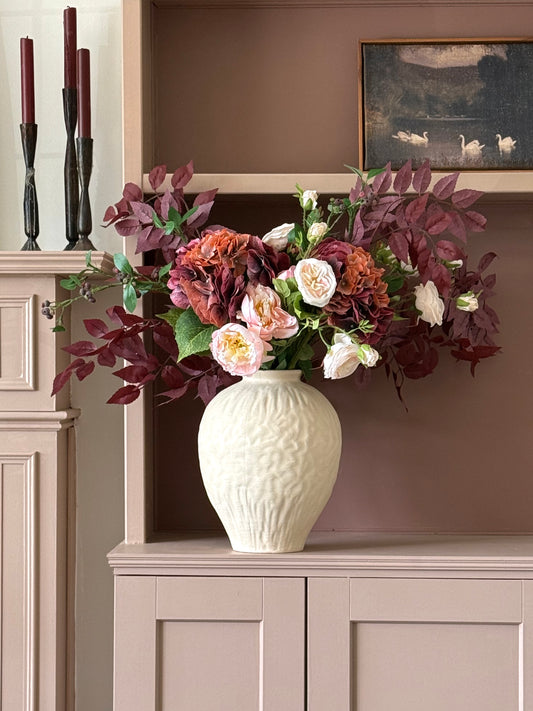 A large artificial floral arrangement in a textured cream ceramic vase, displayed on a dusty pink cabinet against a brown wall.