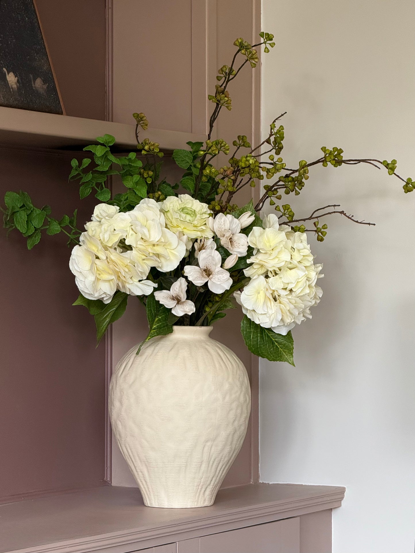 A textured cream-colored ceramic vase filled with a floral arrangement of white hydrangeas, green leaves, and delicate green-budded branches, displayed on a dusty mauve-colored mantelpiece against a neutral wall.