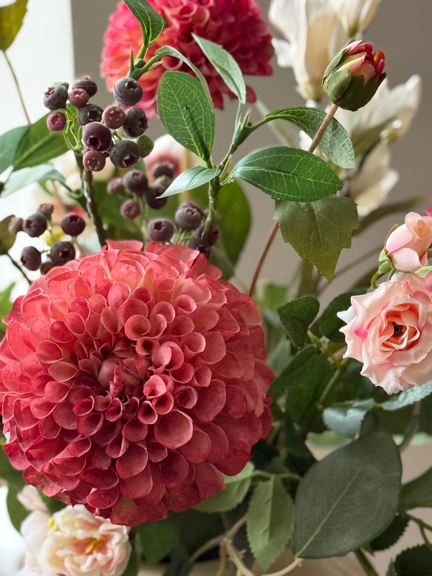 A close-up of a floral arrangement featuring a large coral-pink dahlia, a peach rose, green leaves, and small dark berries against a soft neutral background.
