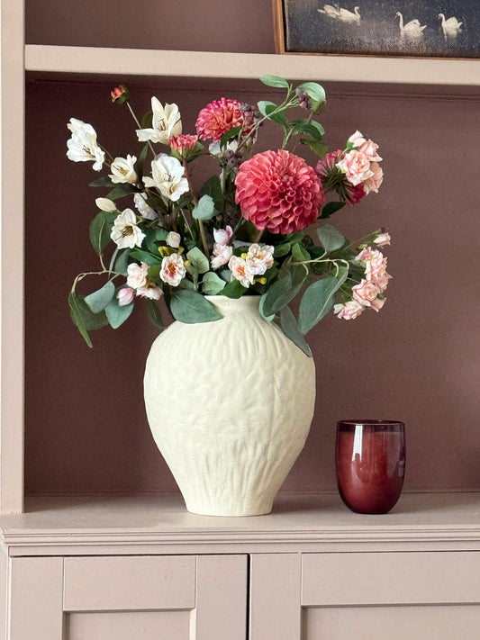 A cream-colored textured ceramic vase filled with a floral arrangement of pink, white, and light pink flowers with green leaves, displayed on a light beige cabinet against a dark brown wall.