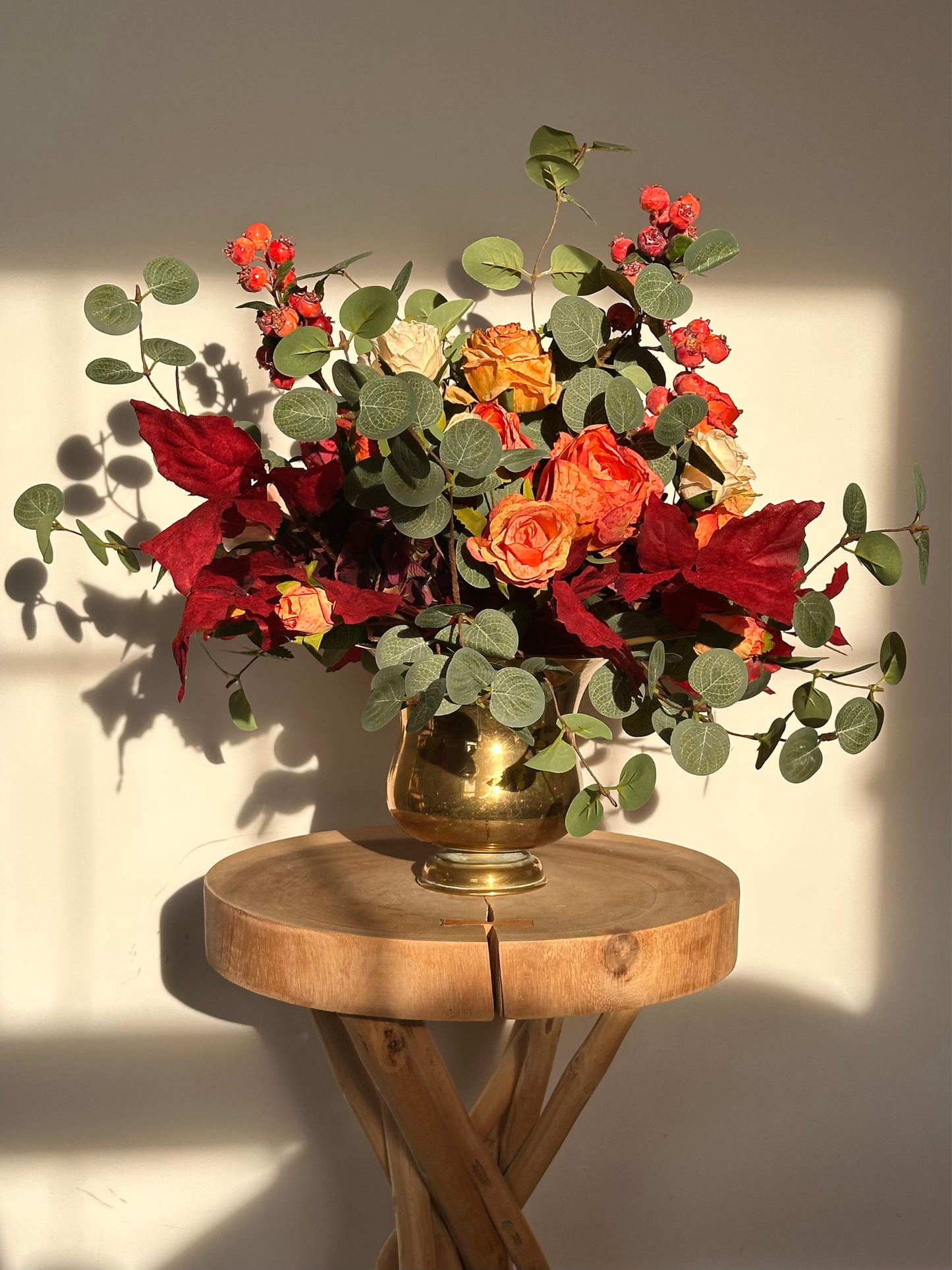 Bouquet of red, orange, and green faux flowers in a gold vase on a wooden table with a neutral background