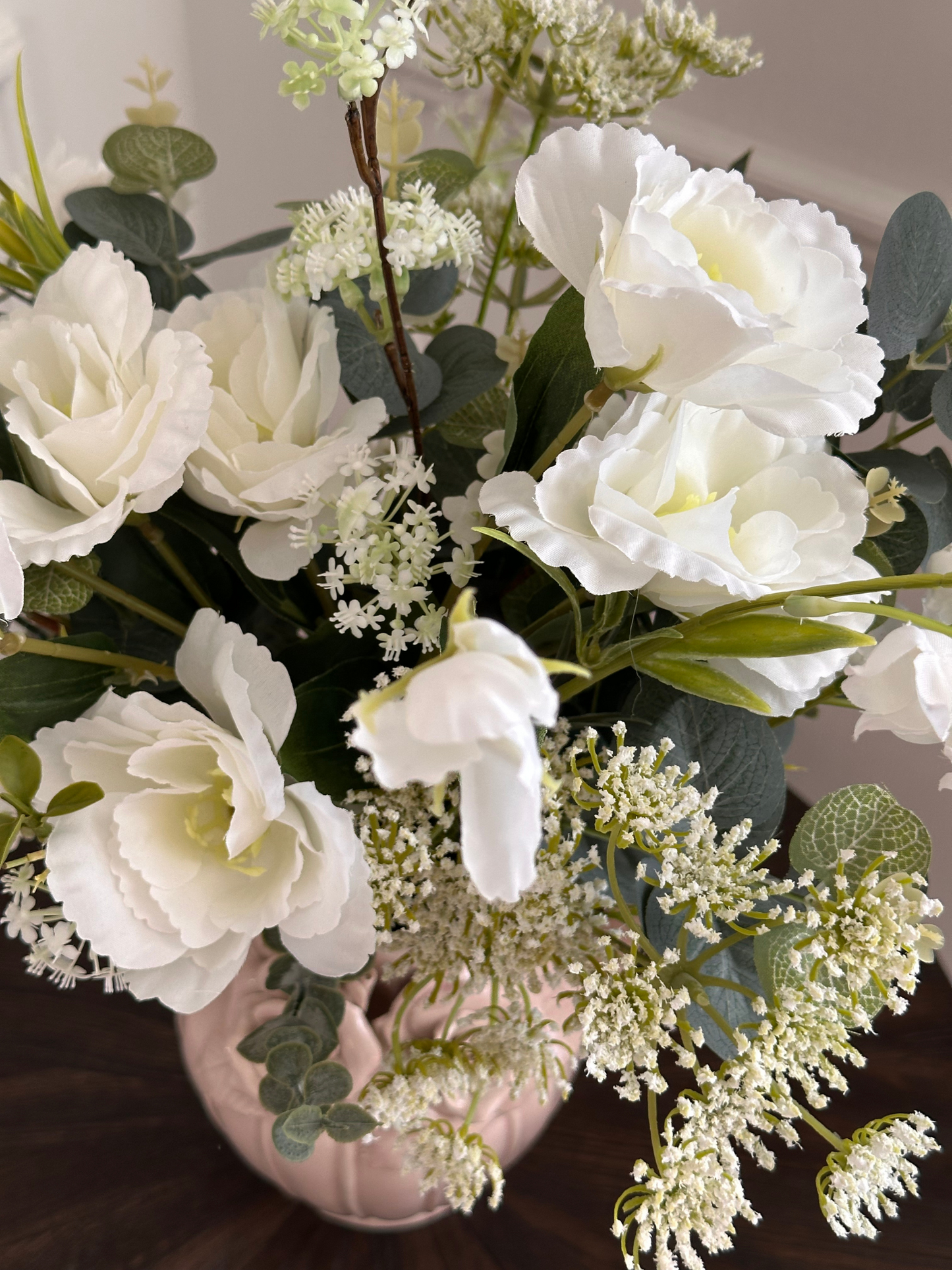 Bouquet of white flowers with green leaves in a pink vase on a dark surface.