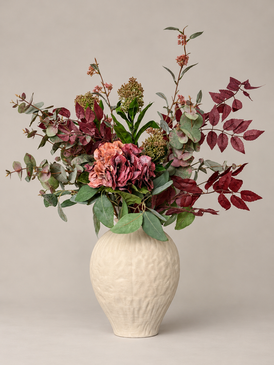 A floral arrangement featuring burgundy and pink flowers, green and silver eucalyptus leaves, and dried botanicals, presented in a textured cream-colored ceramic vase against a neutral beige background.
