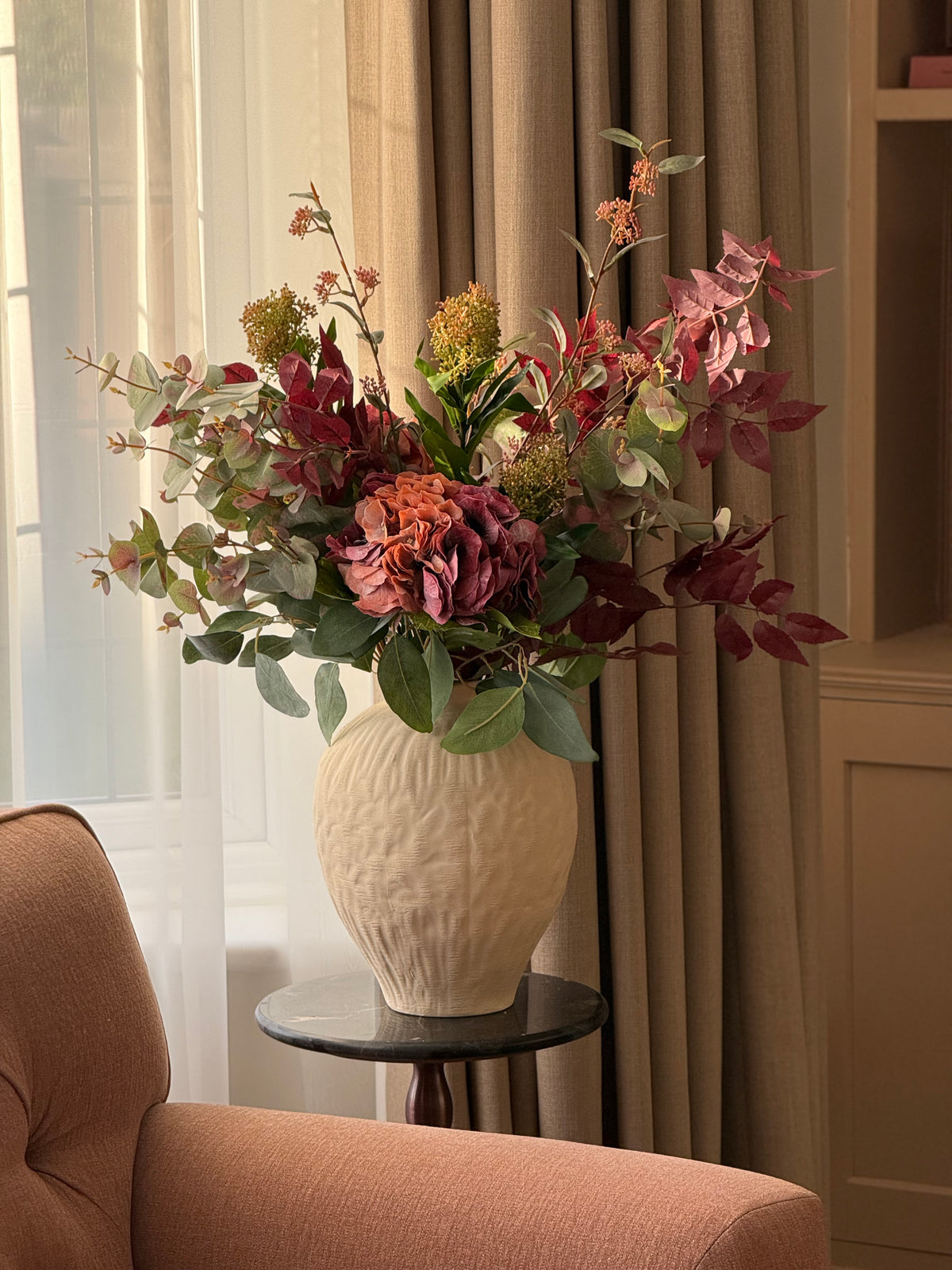 A large textured cream ceramic vase filled with an autumnal floral arrangement of burgundy, dusty pink, and olive green faux flowers, styled on a small black side table next to a peach armchair in a warm living room setting.