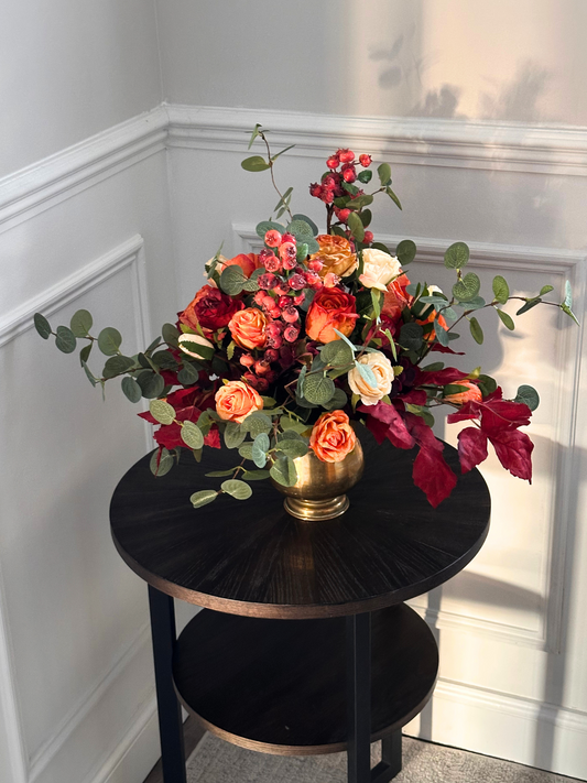 Artificial Floral arrangement in a gold vase on a black round table against a white wall.