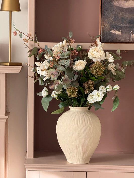 A large cream-colored textured ceramic vase filled with a floral arrangement of white roses, green eucalyptus, and dusty pink filler flowers, displayed on a matching pink shelf next to a gold lamp.