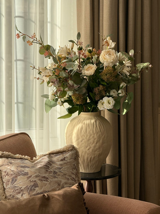 A large cream-colored textured ceramic vase filled with a dense arrangement of white roses, green eucalyptus, and dried botanicals, styled on a side table in a warm, neutral-toned living room next to a brown armchair.