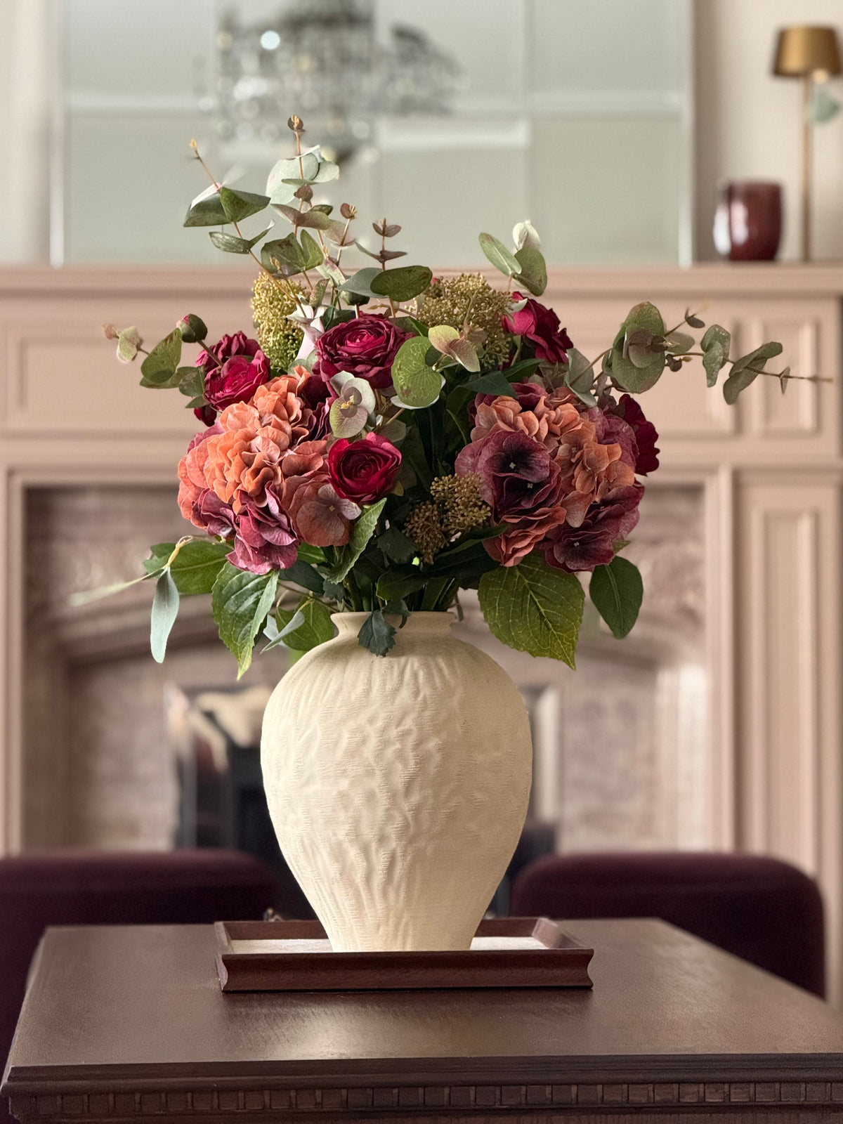 A faux floral arrangement in a textured cream ceramic vase, placed on a dark wooden table in front of a softly blurred fireplace background.