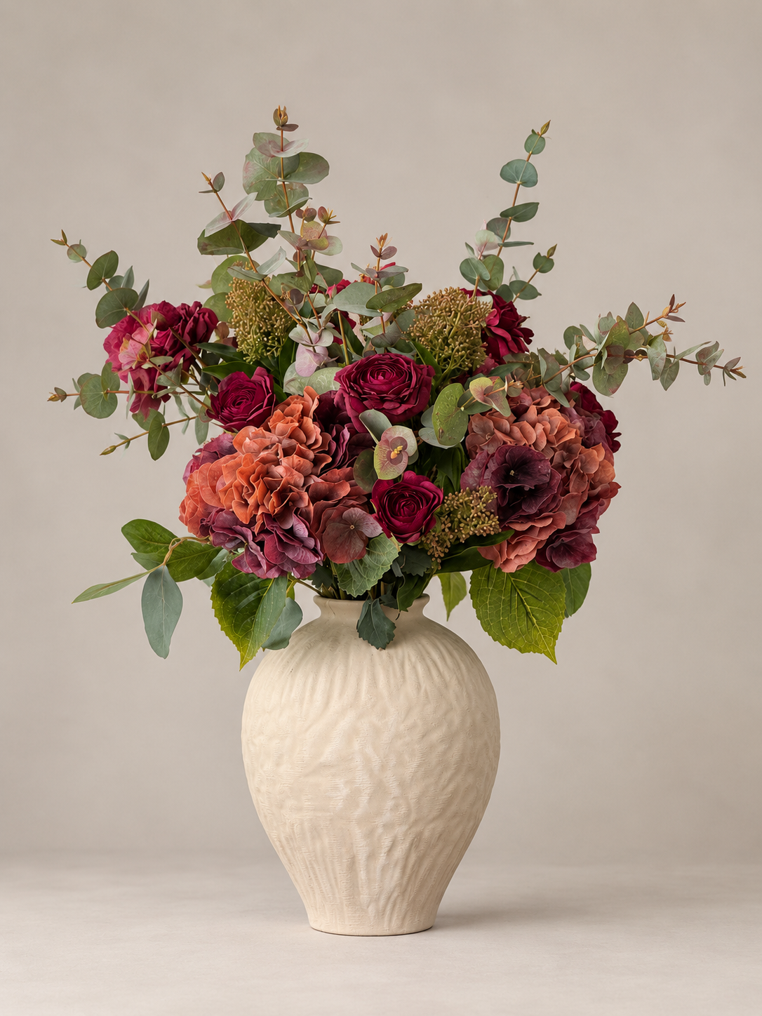 A floral arrangement featuring burgundy roses, mauve hydrangeas, and green eucalyptus stems in a textured cream-colored ceramic vase, set against a neutral beige background.