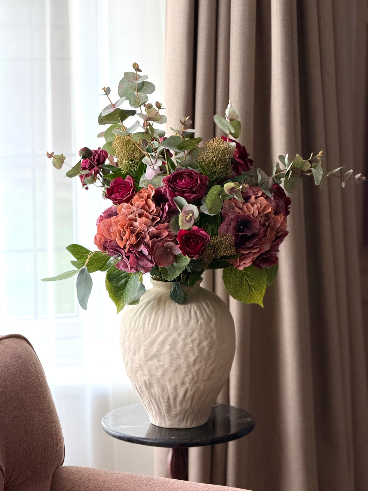A faux floral arrangement in a textured cream ceramic vase, displayed on a small round side table in a living room setting with beige curtains and a sofa.