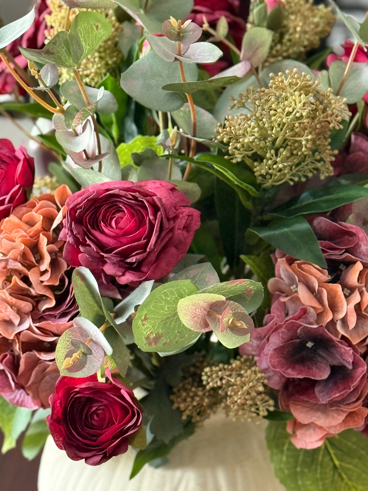 A close-up of a floral arrangement featuring deep burgundy roses, dusty pink hydrangeas, sage green eucalyptus, and textured beige flower clusters, set against a soft beige background.