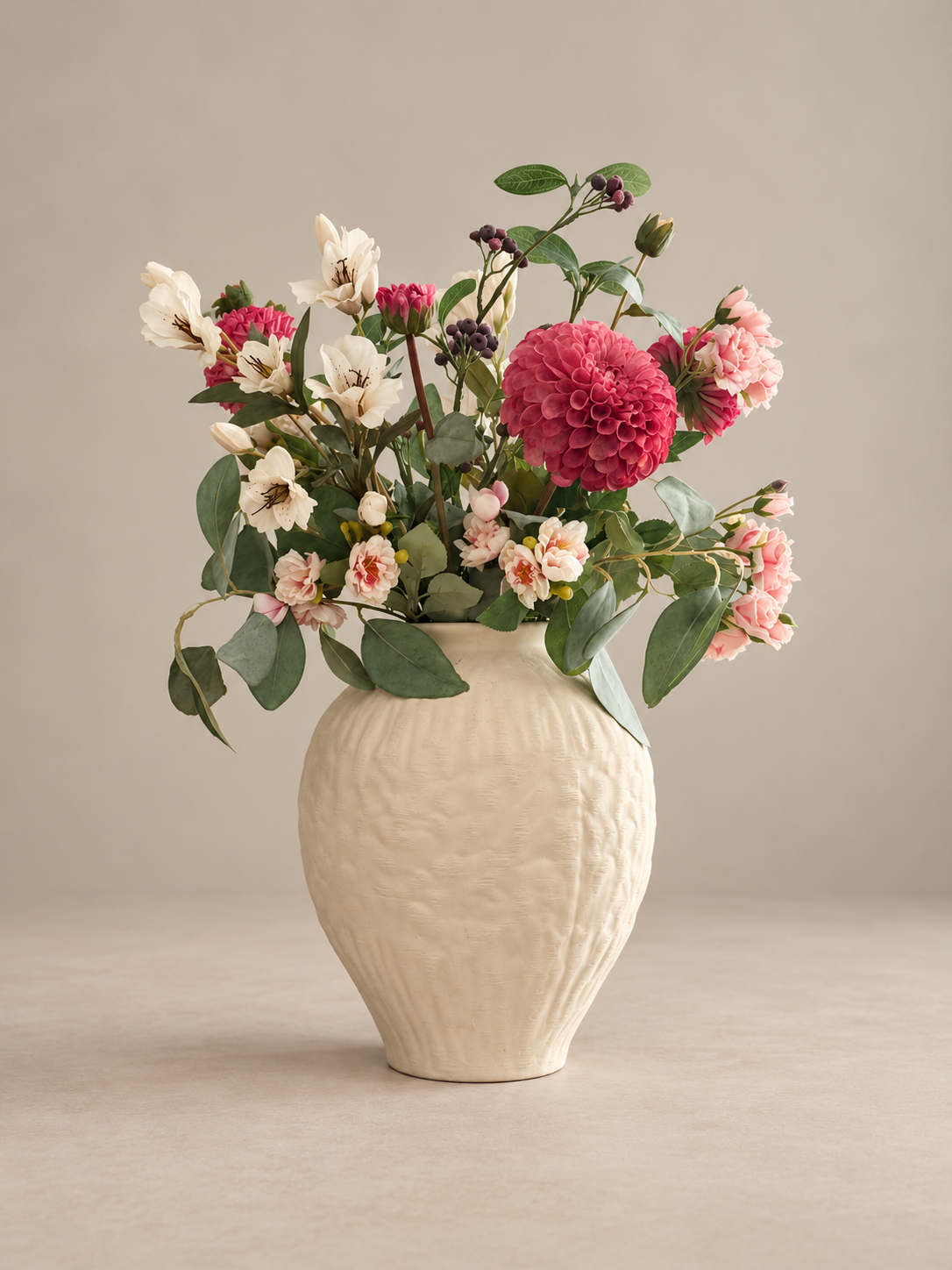 A cream-colored textured ceramic vase filled with a colorful floral arrangement featuring pink, red, and white flowers with green leaves, set against a neutral beige background.