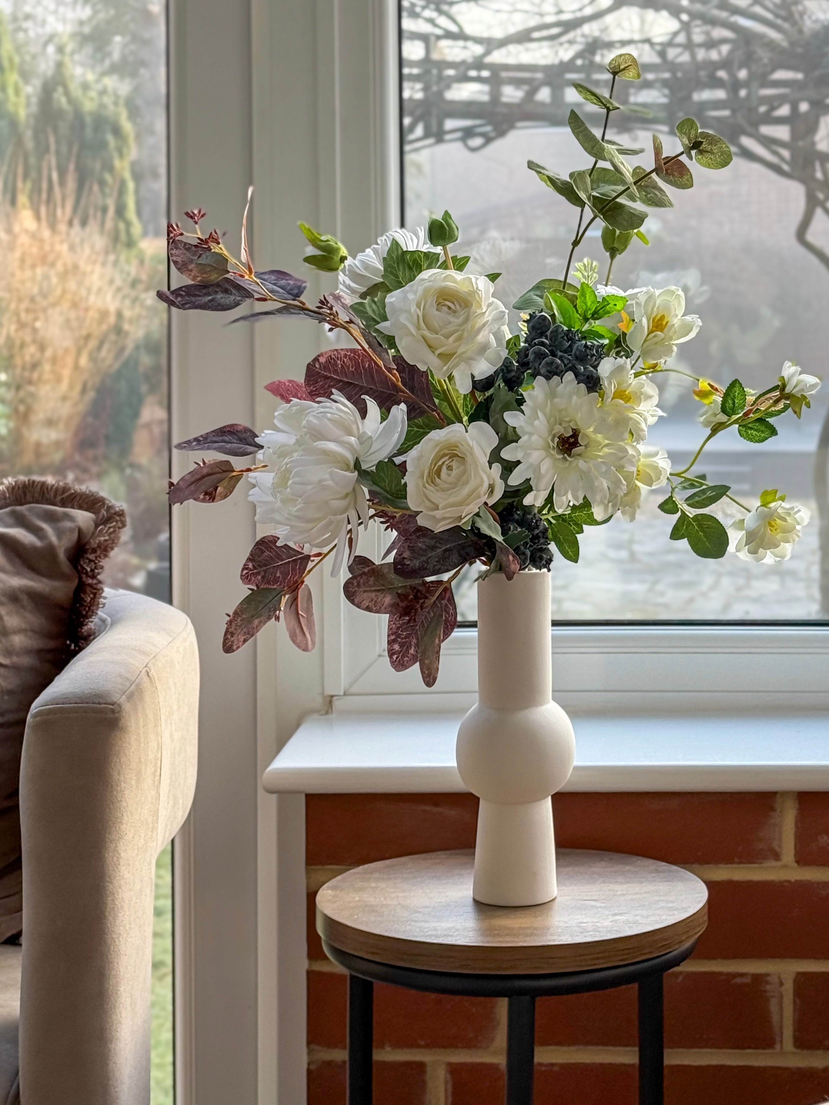 Artificial Floral arrangement in a white vase on a small round table by a window.
