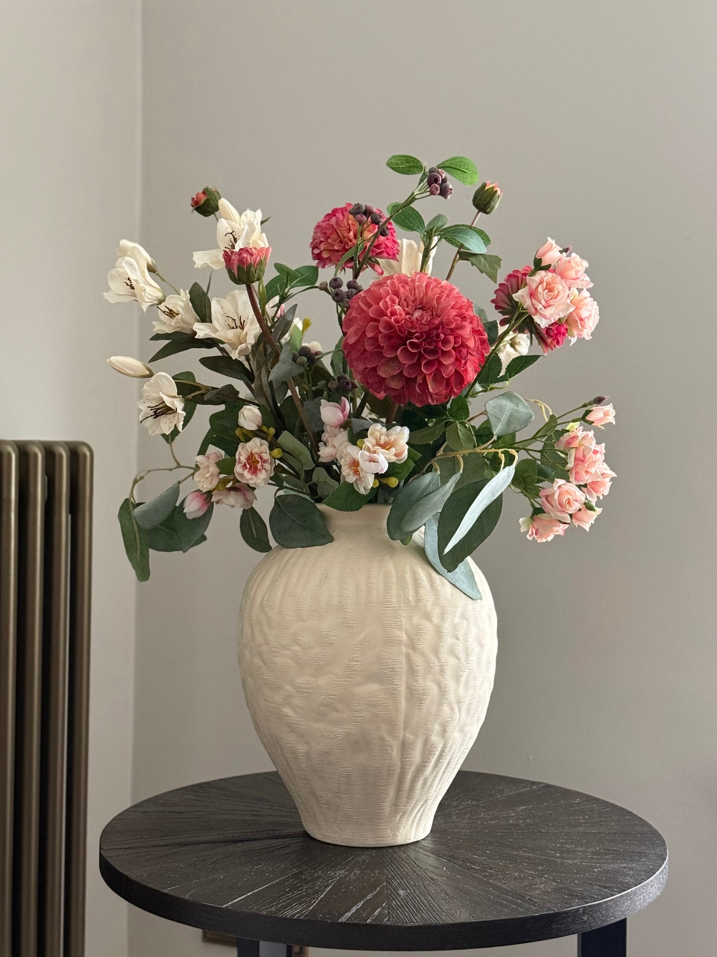 A textured cream ceramic vase filled with a colorful floral arrangement featuring a large pink dahlias, pink roses, white lilies, and green foliage, displayed on a round black table against a neutral gray wall.