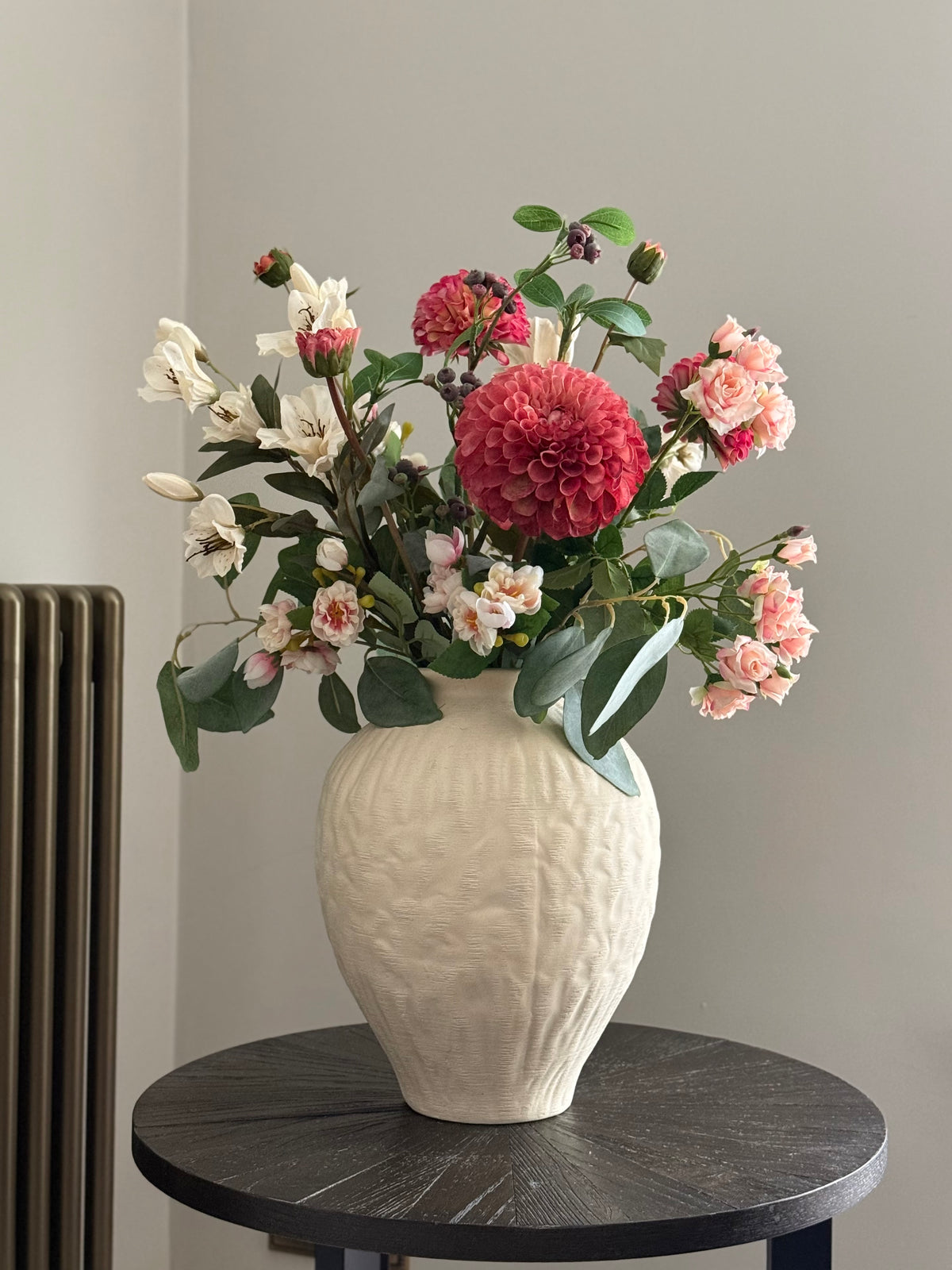 A textured cream ceramic vase filled with a colorful floral arrangement featuring a large pink dahlias, pink roses, white lilies, and green foliage, displayed on a round black table against a neutral gray wall.
