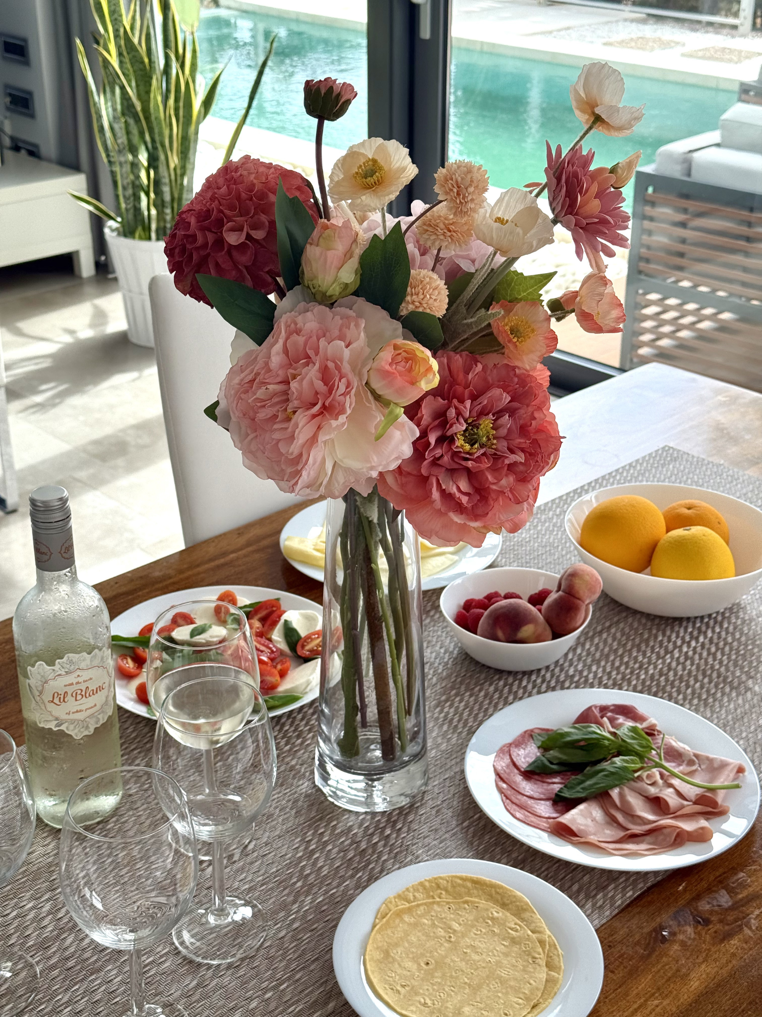 Dining table set with artificial flowers, food, and drinks in a bright room.