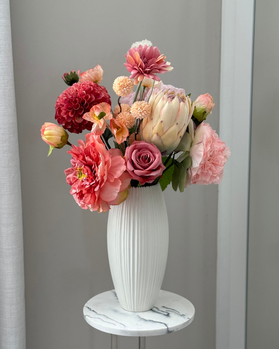 White vase with colourful Best & Bloom artificial flowers on a marble shelf against a gray curtain background
