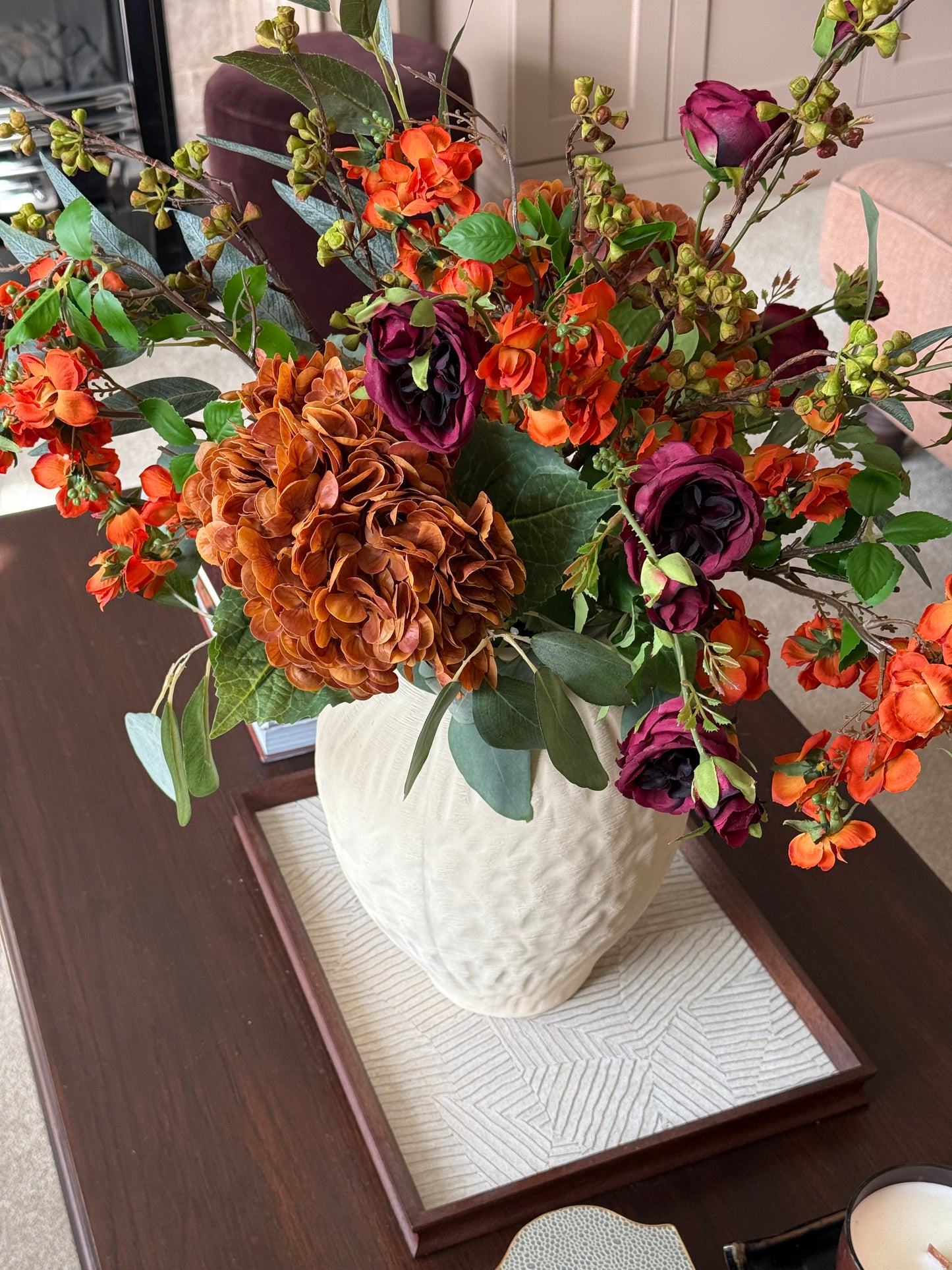An autumn floral arrangement in a textured white ceramic vase, placed on a wooden tray on a dark wood table. The bouquet features orange hydrangea-like blooms, orange roses, dark burgundy roses, and green foliage.