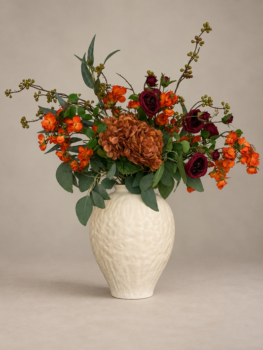 An autumn floral arrangement in a textured cream ceramic vase, featuring orange blossoms, burgundy roses, and brown hydrangea-like flowers with green foliage, set against a neutral beige background.