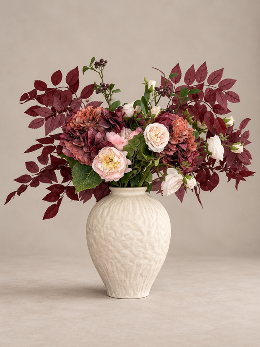 A floral arrangement featuring burgundy faux leaves, pink and white roses, and green foliage in a textured cream-colored ceramic vase, set against a neutral beige background.