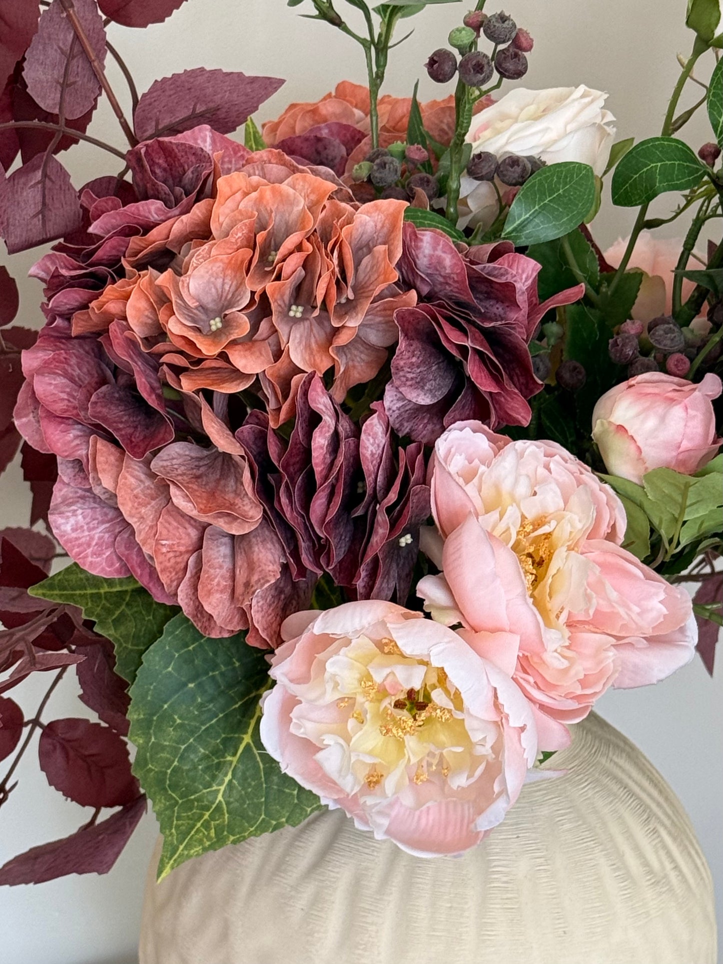 A close-up of a floral arrangement featuring large ruffled hydrangea blooms in dusty rose and mauve, pale pink peony-like flowers, green leaves, and burgundy berries, set in a cream-colored vase against a light neutral background.