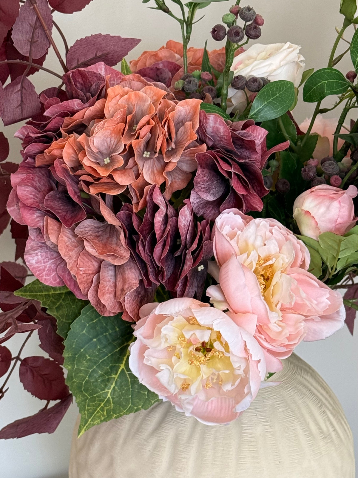 A close-up of a floral arrangement featuring large ruffled hydrangea blooms in dusty rose and mauve, pale pink peony-like flowers, green leaves, and burgundy berries, set in a cream-colored vase against a light neutral background.