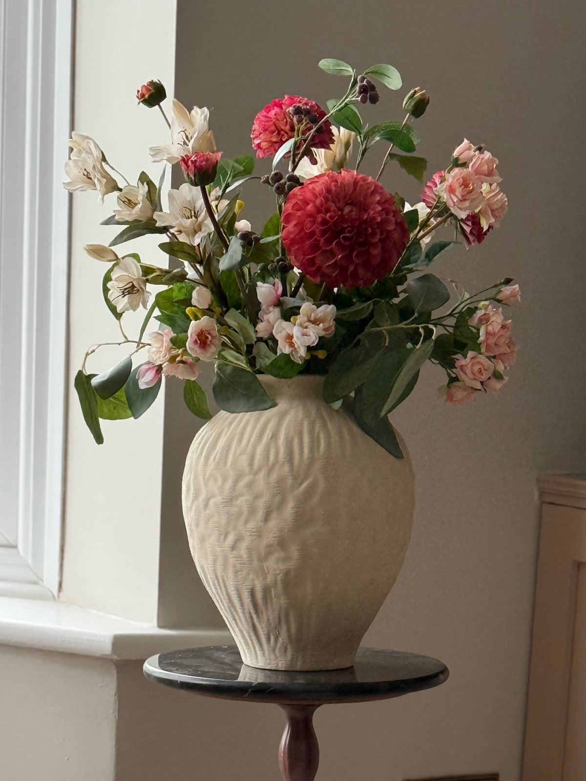 A ceramic vase with a textured, ribbed cream finish holding a vibrant floral arrangement of pink, red, and white flowers with green leaves, displayed on a dark round side table next to a window.