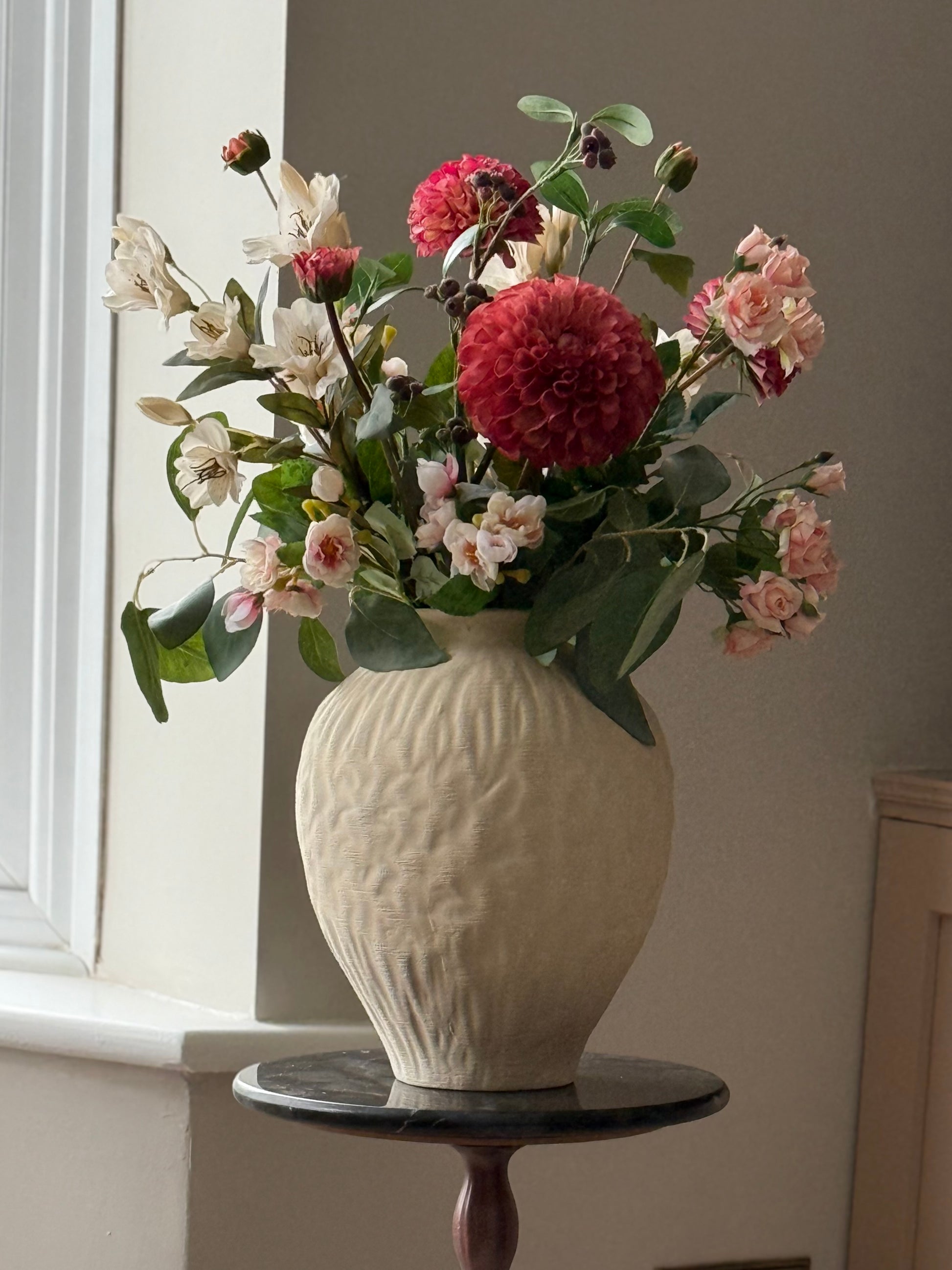 A ceramic vase with a textured, ribbed cream finish holding a vibrant floral arrangement of pink, red, and white flowers with green leaves, displayed on a dark round side table next to a window.