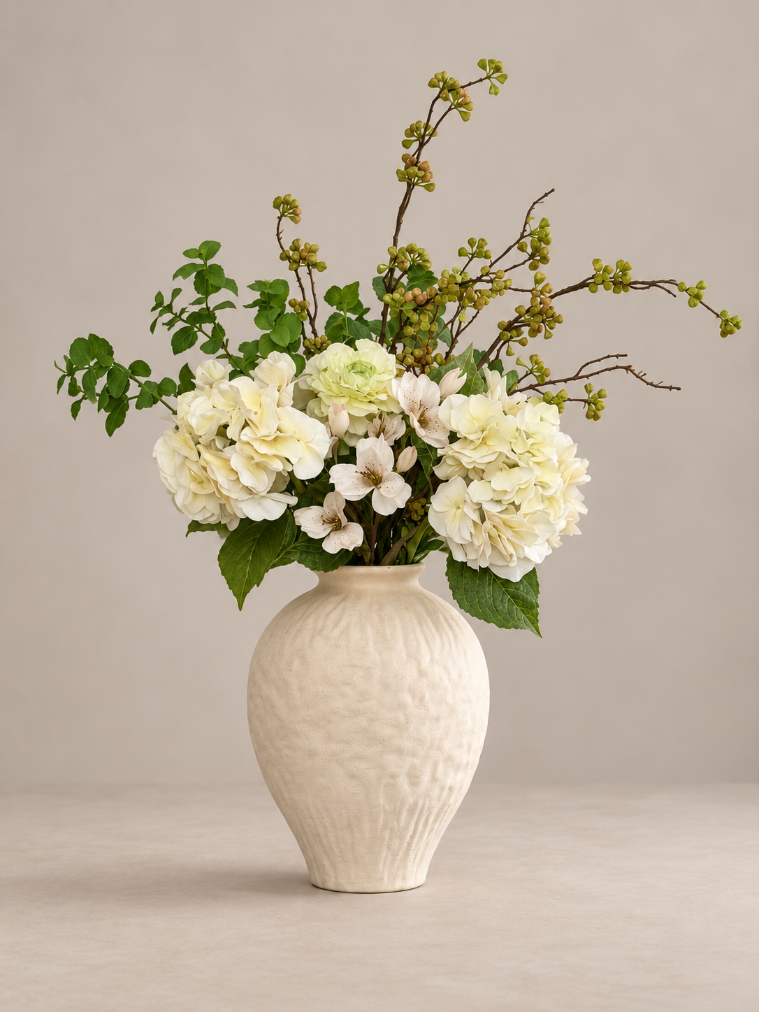 A floral arrangement featuring cream and white flowers with green foliage, displayed in a textured cream ceramic vase against a neutral beige background.