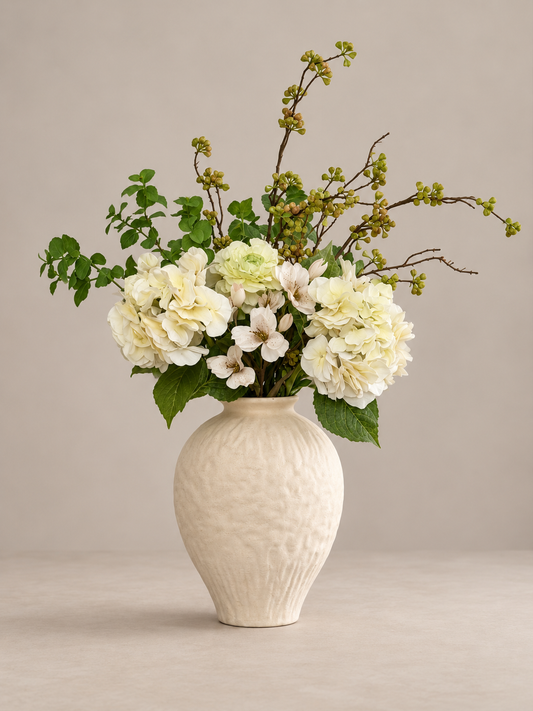 A floral arrangement featuring cream and white flowers with green foliage, displayed in a textured cream ceramic vase against a neutral beige background.
