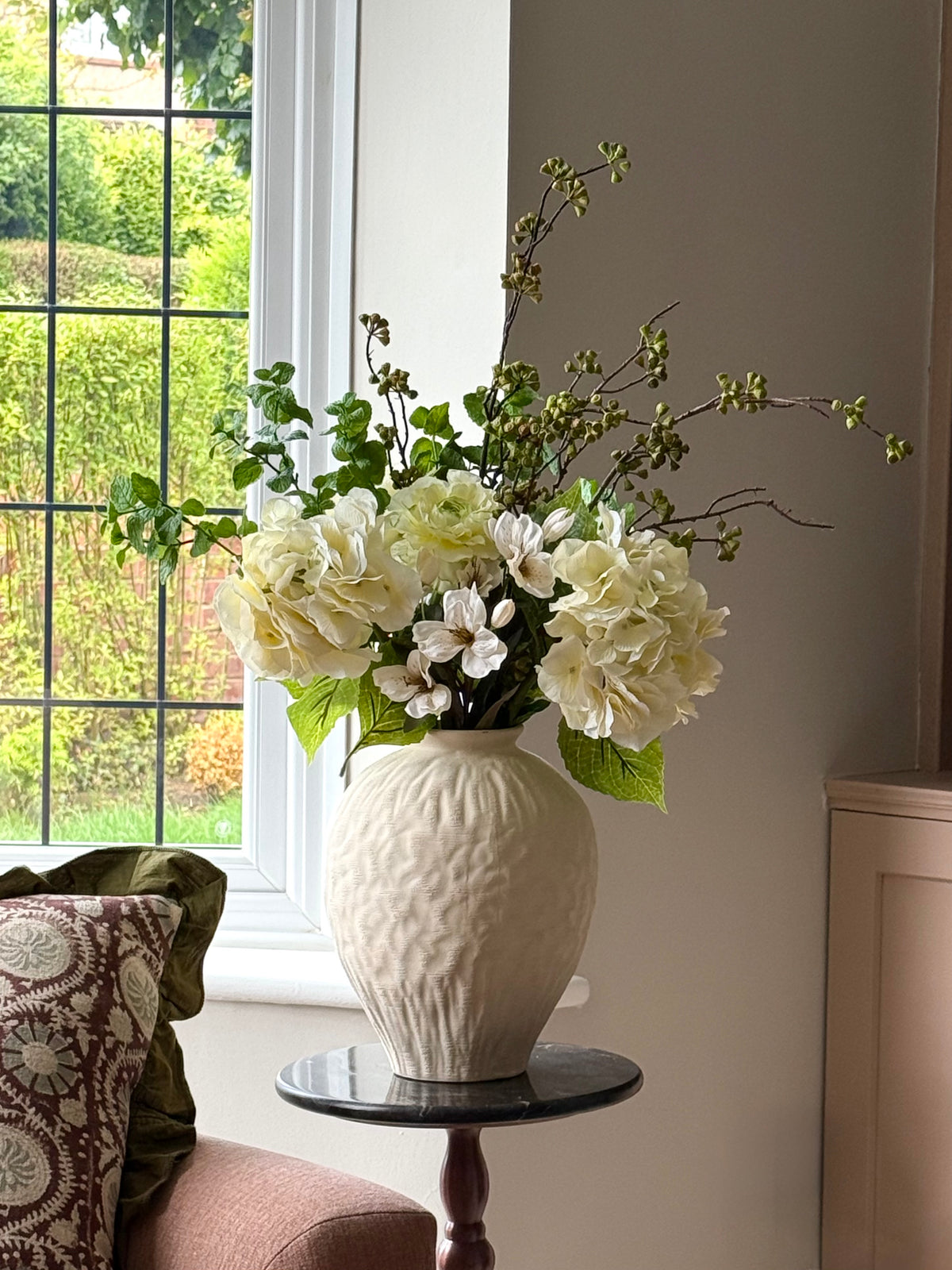 A cream-colored textured ceramic vase filled with white and cream artificial flowers and green foliage, displayed on a small dark marble-topped side table in a living room setting next to a window.