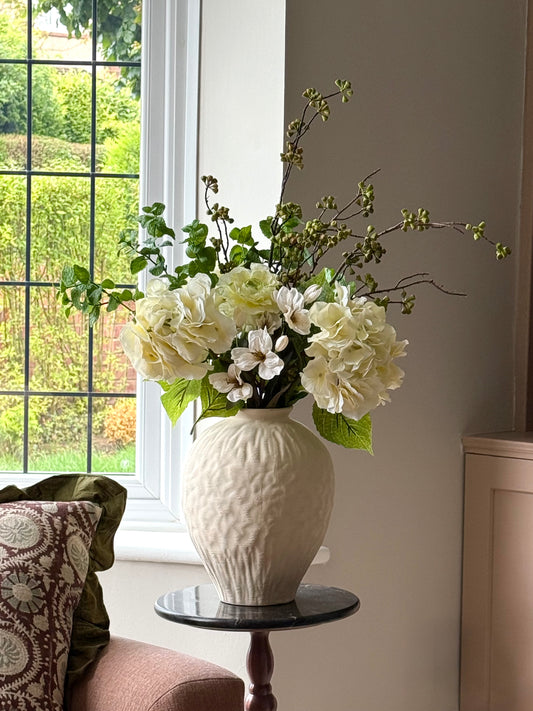 A cream-colored textured ceramic vase filled with white and cream artificial flowers and green foliage, displayed on a small dark marble-topped side table in a living room setting next to a window.