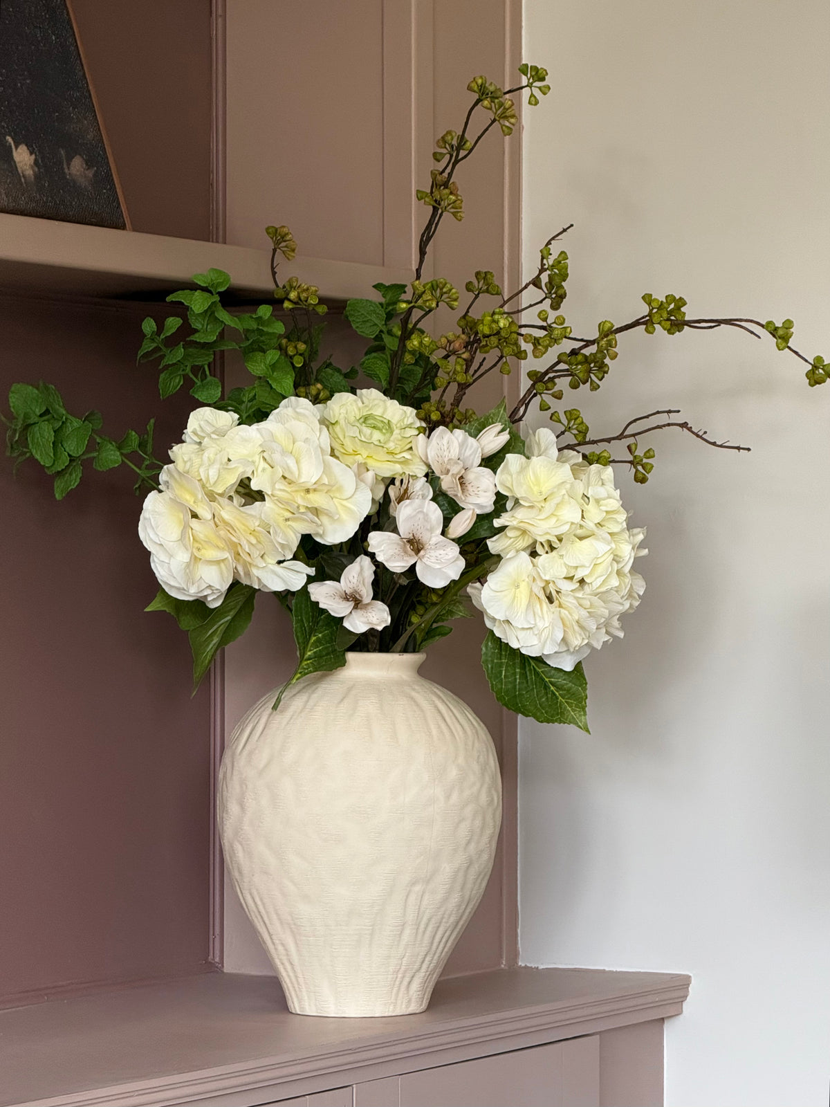 A textured cream-colored ceramic vase filled with a floral arrangement of white hydrangeas, green leaves, and delicate green-budded branches, displayed on a dusty mauve-colored mantelpiece against a neutral wall.