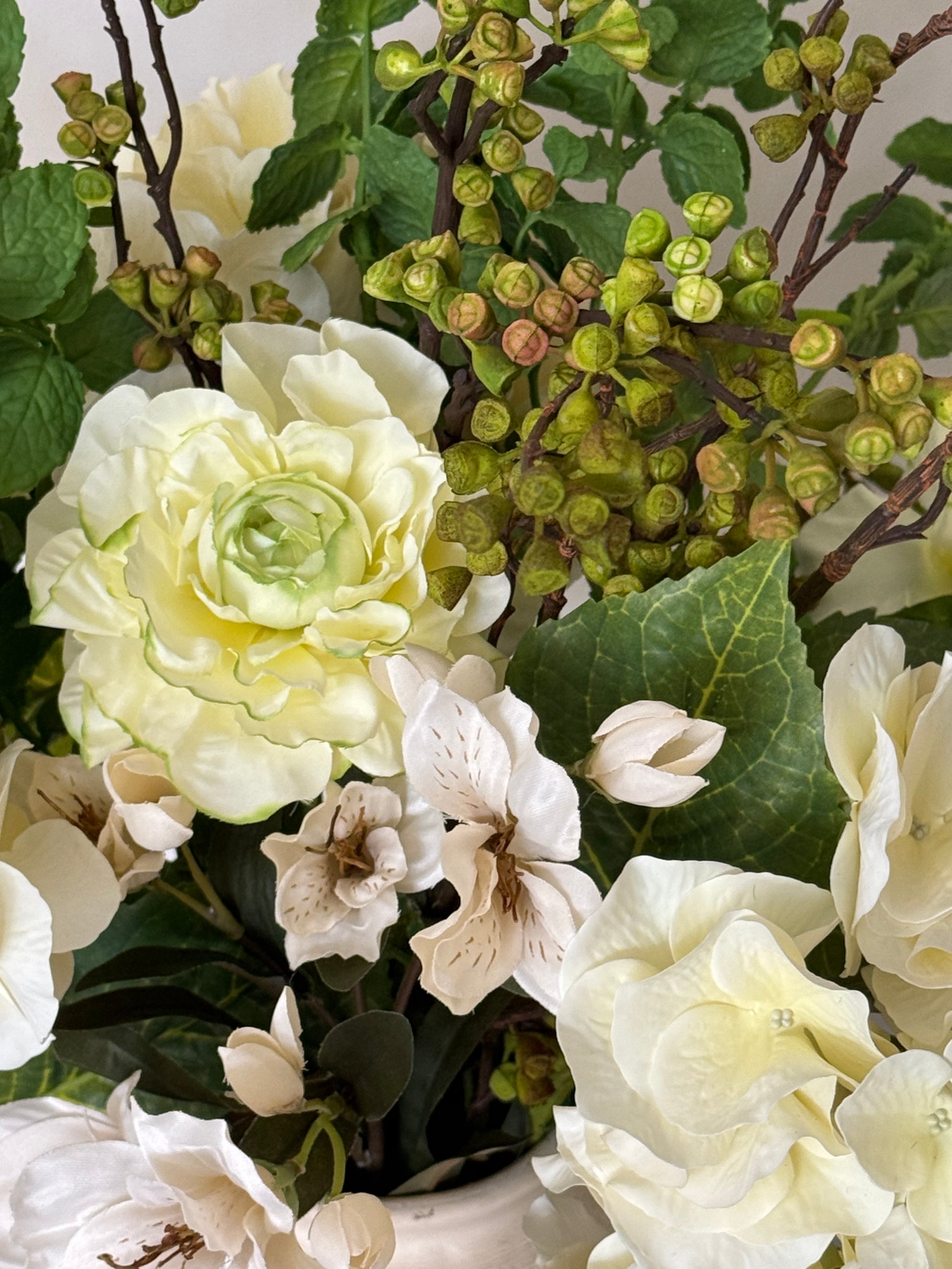 A close-up of an artificial floral arrangement featuring cream-colored roses, white lilies, green leaves, and clusters of small green buds on brown stems.
