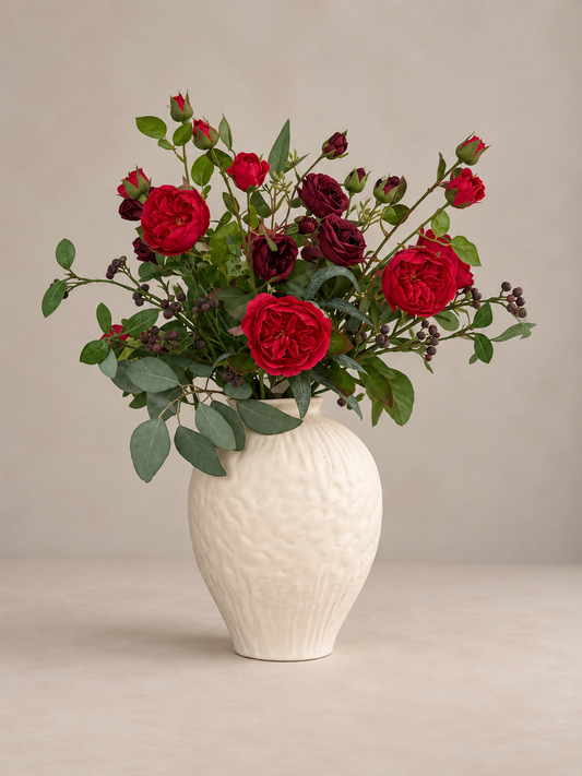 A floral arrangement of red and deep burgundy roses with green foliage, presented in a textured off-white ceramic vase against a neutral beige background.