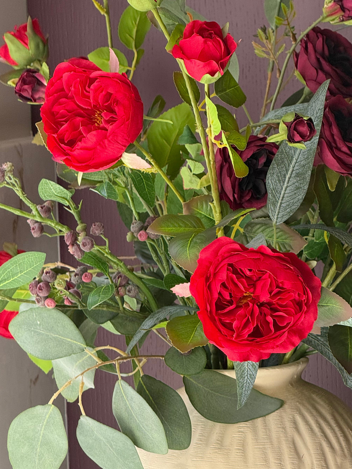 A bouquet of artificial roses in deep red and dark burgundy tones with green leaves, arranged in a textured cream-colored ceramic vase against a dark purple wall.
