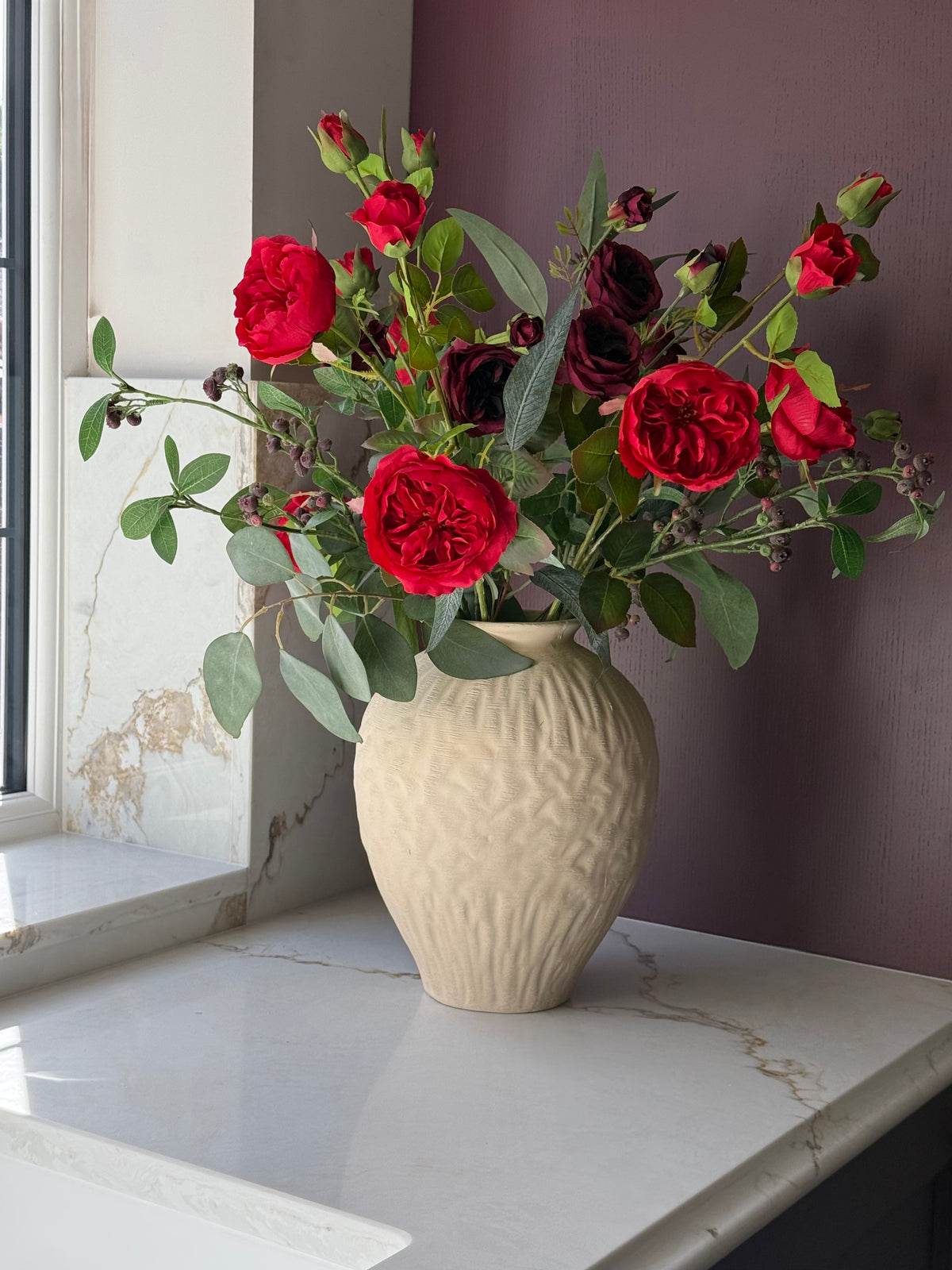 A faux rose floral arrangement in a textured cream ceramic vase, set on a white marble windowsill with a dark purple wall in the background.