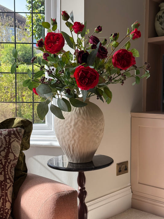 A large cream-colored textured ceramic vase filled with a bouquet of red and dark burgundy roses with green leaves, displayed on a small black-topped side table in a sunlit room next to a window.