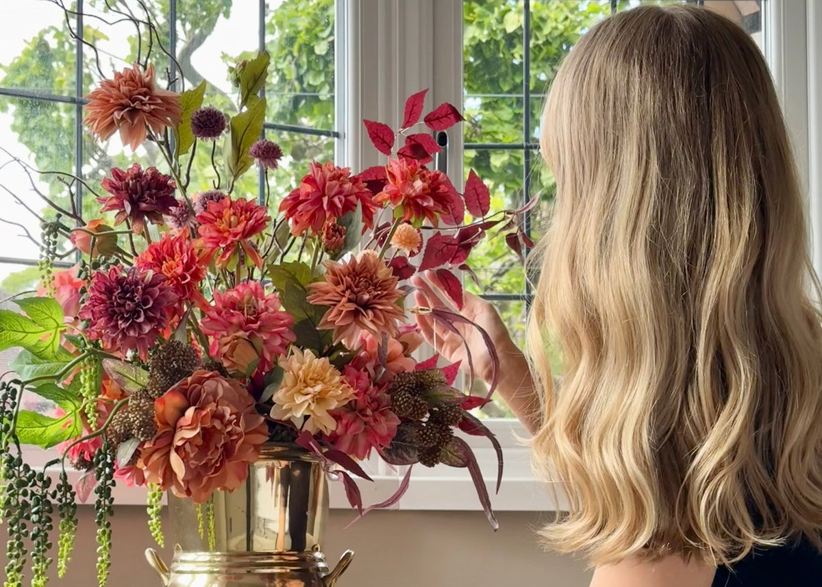 A woman with blonde hair sits at a table by a window, holding a large bronze vase filled with a colorful bouquet of red, pink, and orange flowers.
