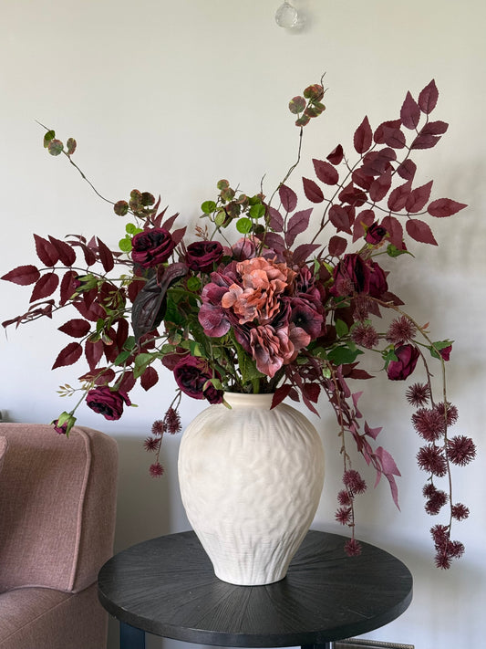 White textured vase with a large artificial floral arrangement on a dark round table against a white wall.