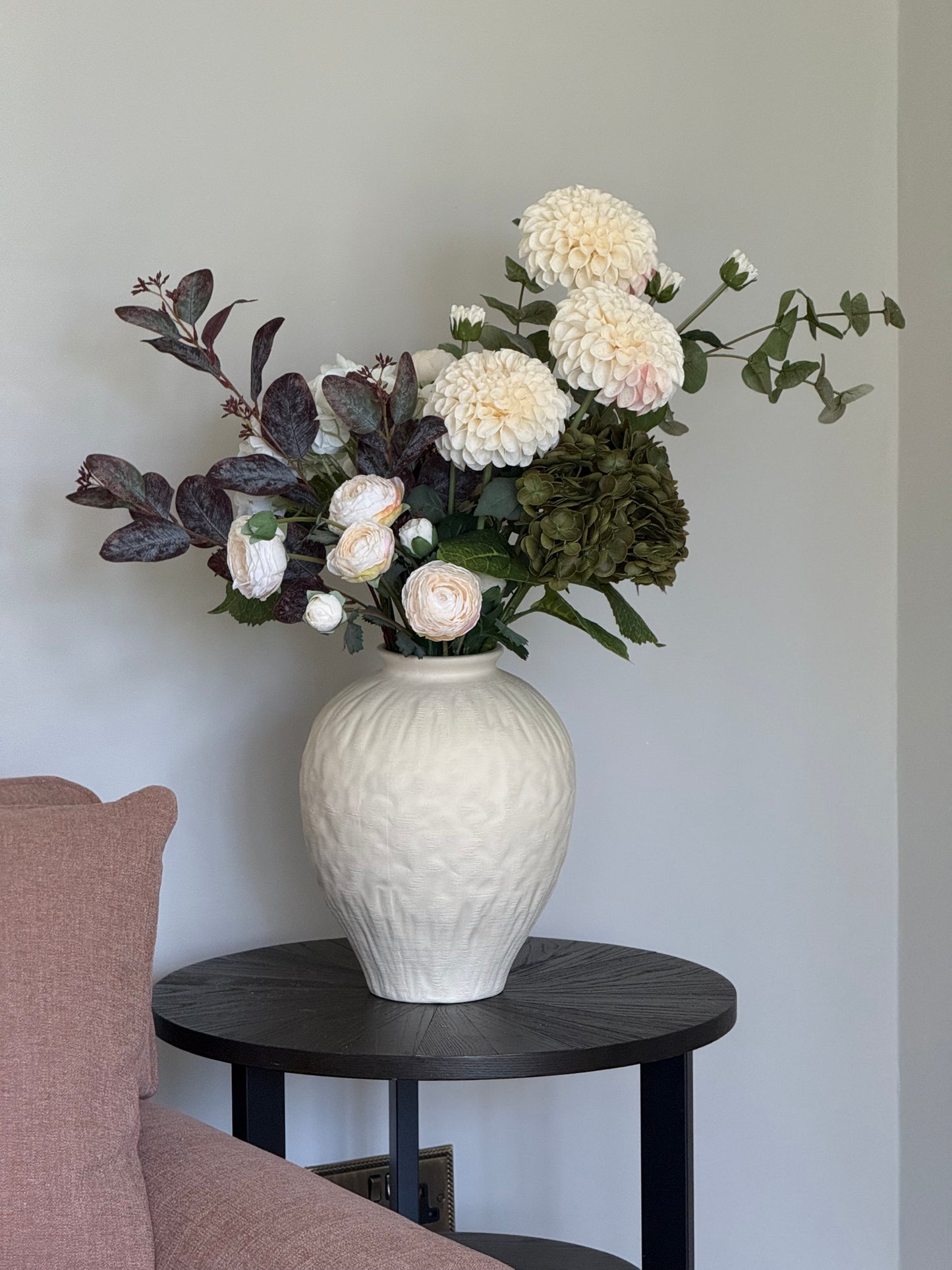 White textured vase with flowers on a black side table against a light gray wall.