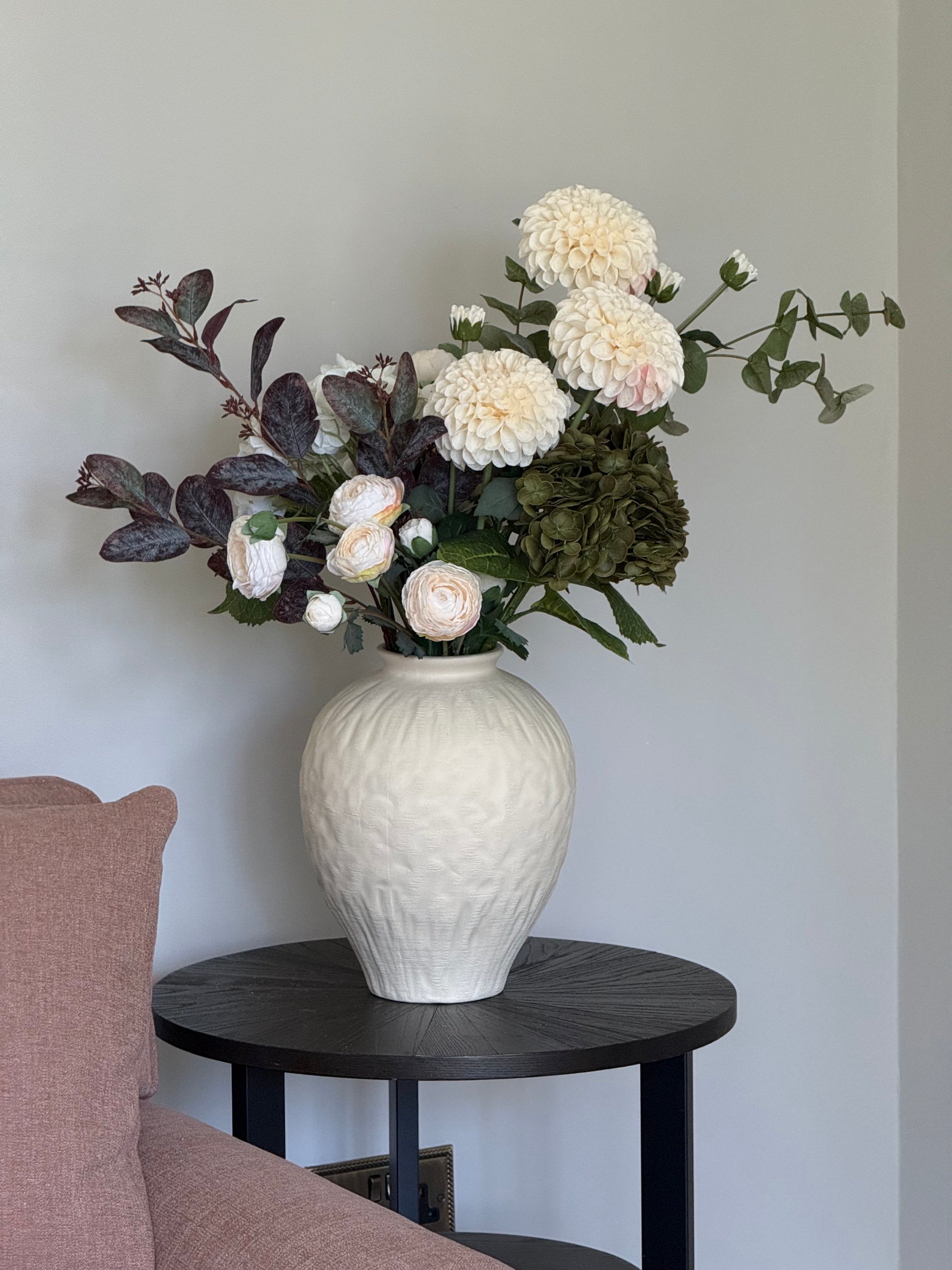 White textured vase with flowers on a black side table against a light gray wall.