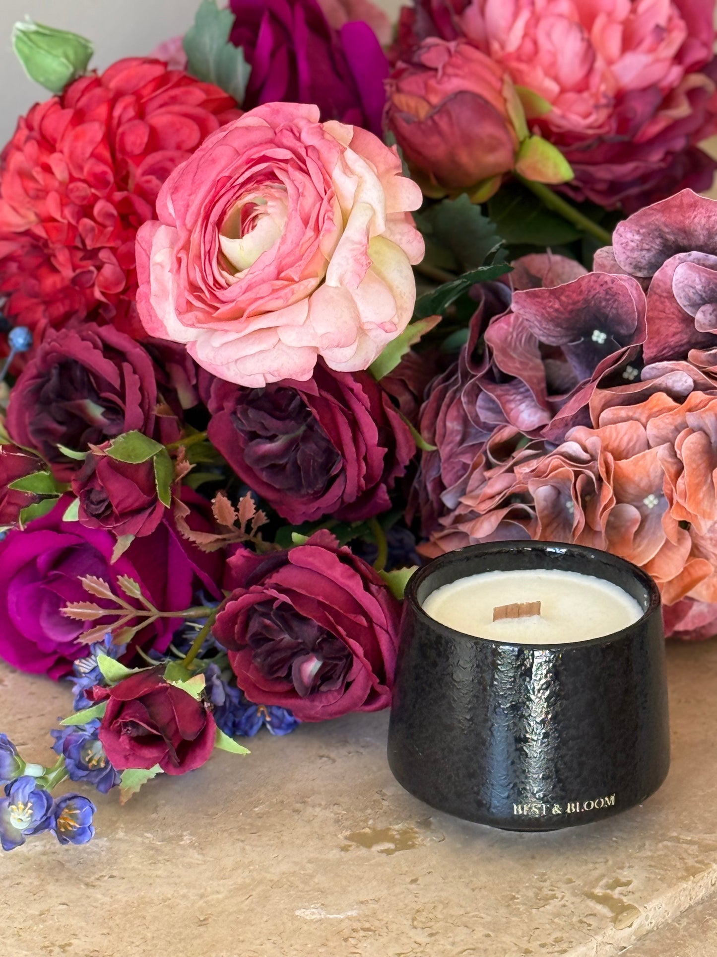 Black candle with 'Best & Bloom' branding in front of a bouquet of colorful flowers on a stone surface.