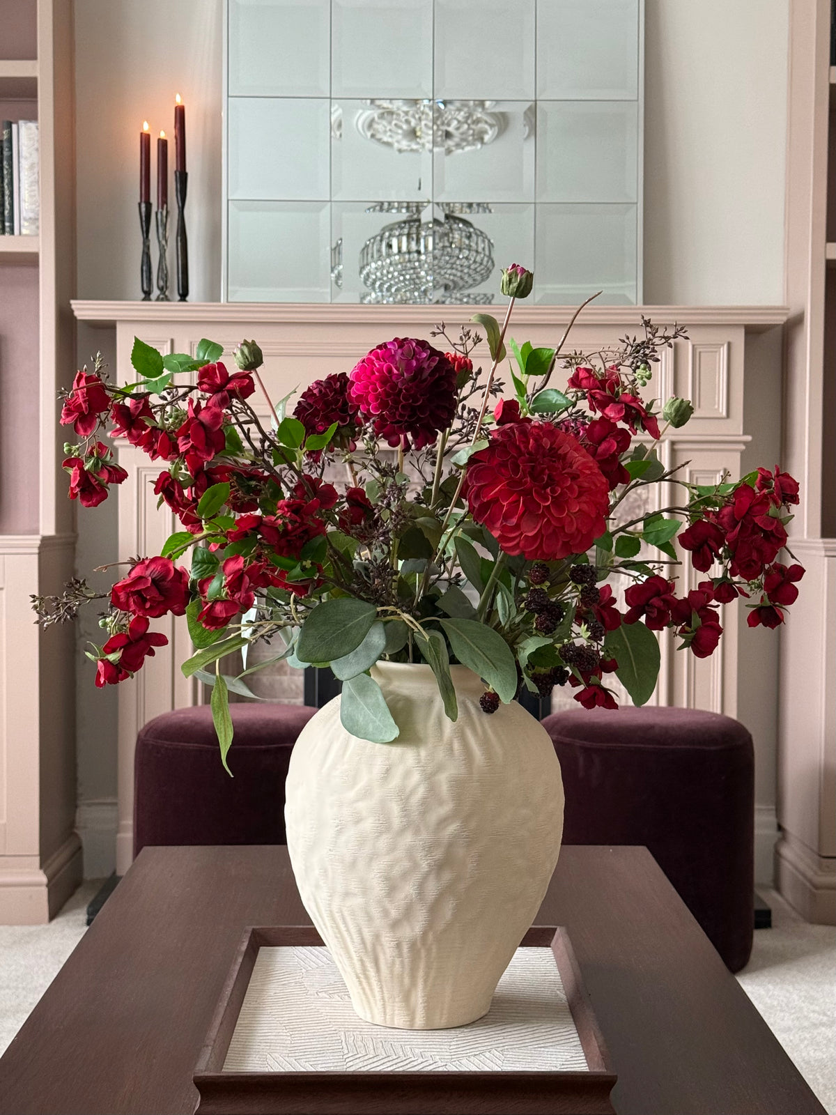 A large textured cream ceramic vase filled with a lush arrangement of deep red roses and green foliage, displayed on a dark wood table in a cozy, elegant living room setting.