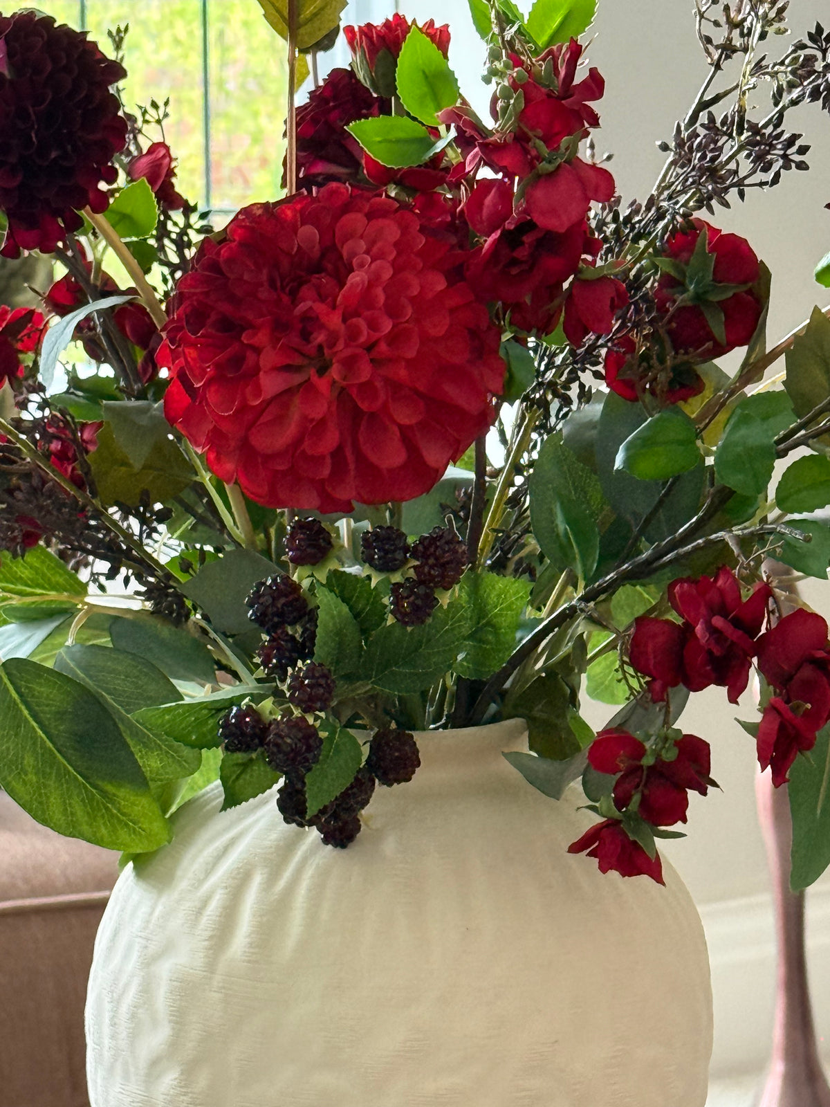 A close-up of a floral arrangement featuring a large red dahlia, smaller red blossoms, green leaves, and dark berry accents, presented in a textured off-white ceramic vase.
