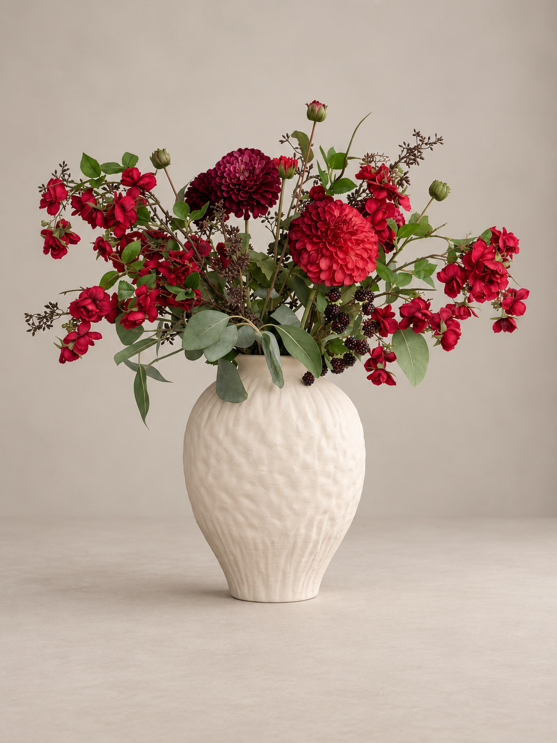 A floral arrangement of deep red flowers and green foliage in a textured off-white ceramic vase, set against a neutral beige background.