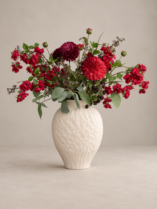 A floral arrangement of deep red flowers and green foliage in a textured off-white ceramic vase, set against a neutral beige background.